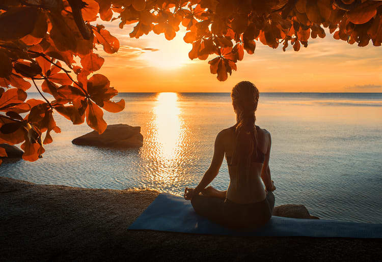 Woman meditating, facing the sun as it sets over the water.