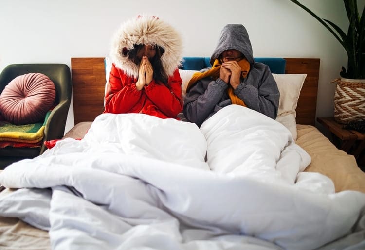 Couple sitting in bed, wearing thick winter jackets.