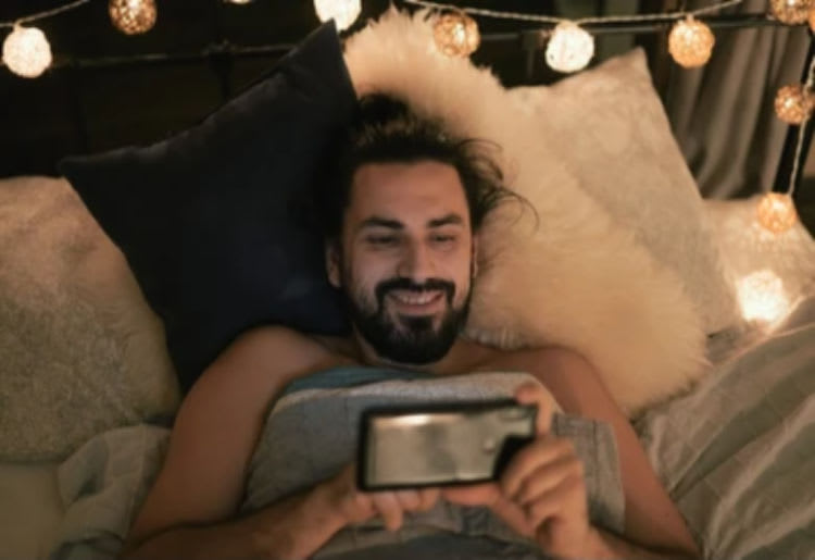 A man laying in bed, smiling as he types something on his phone.