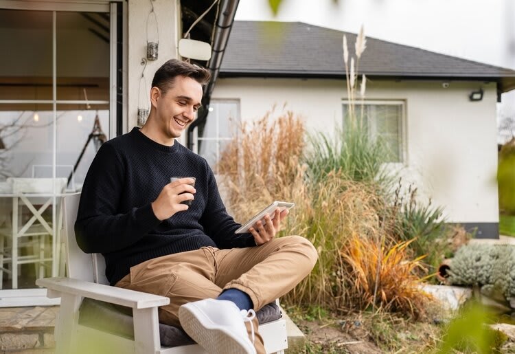 A young man seated on the porch in the backyard of a modern home, smiling while looking at his phone.