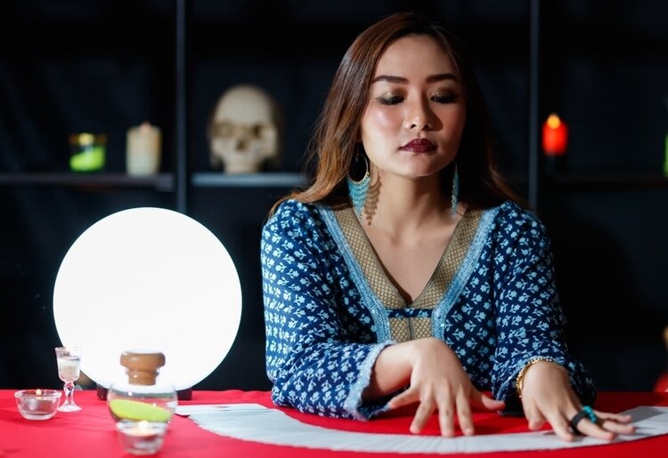 A fortune teller seated next to a glowing crystal ball, with her hands hovering over a sprawled out deck of tarot cards.