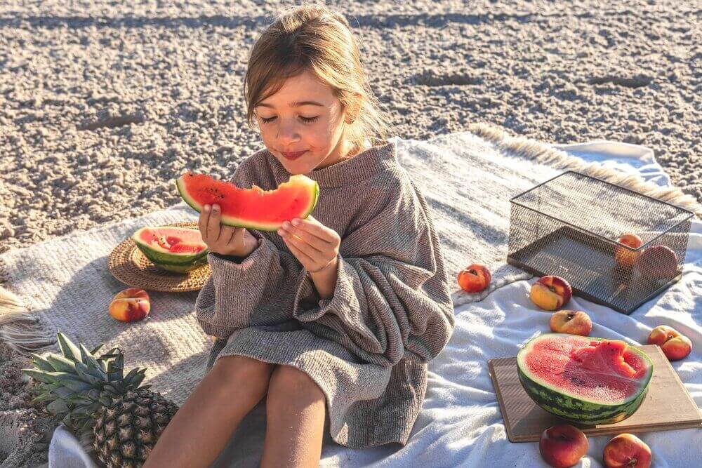 A child seated on the beach, eating watermelon, with various other fruits visible on the towel.
