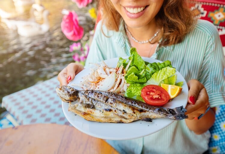 A young adolescent woman smiling whilst holding up a plate of food containing grilled fish and two fresh sides.