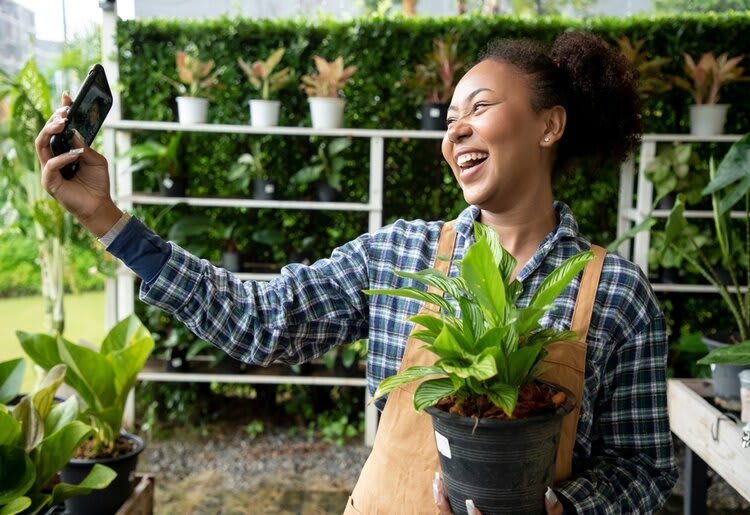 A young nursery assistant taking a selfie with a plant in her hand, and multiple others in the background.