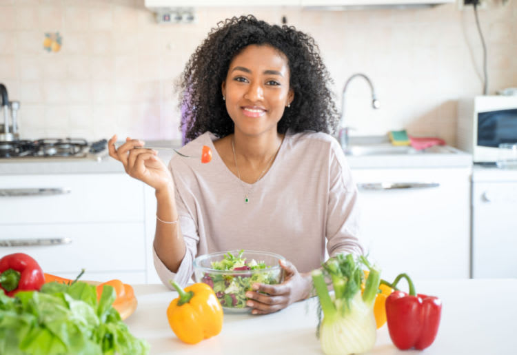 A woman smiling as she eats a bowl of salad, with various vegetables visible on the kitchen counter.