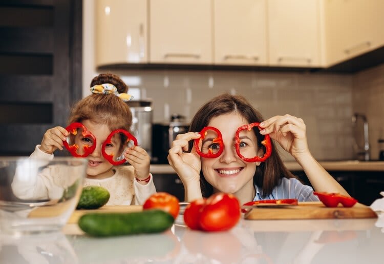 A mother and daughter having fun in the kitchen, holding slices of red pepper around there eyes.