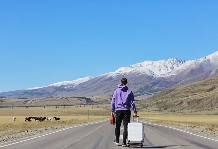 A man standing in the middle of a long open road with his suitcase, facing snow-topped mountains far away.