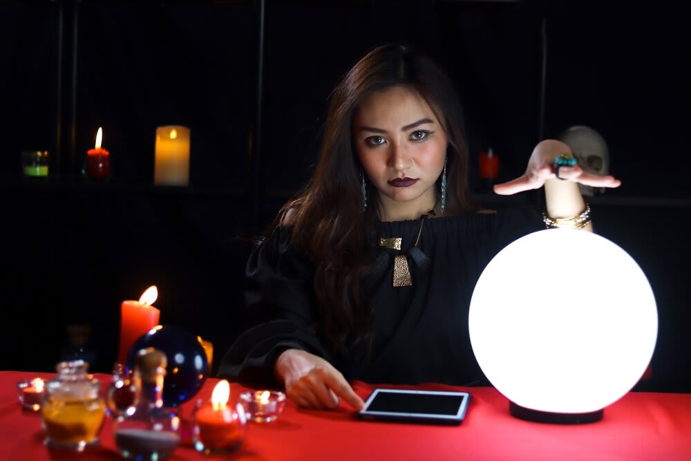 A young psychic medium seated in a dark room with her hand hovering above a glowing crystal ball, with an electronic device, candles, and runes in front of her on a red table.