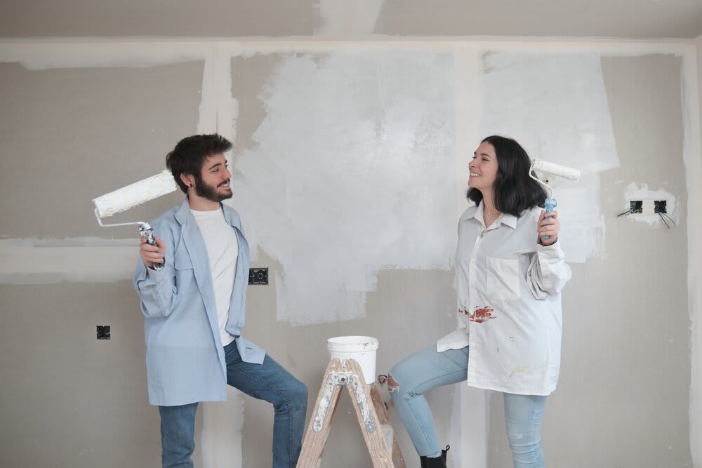 A young couple smiling at one another, each holding a paint roller in their hand, with a portion of the wall already painted.