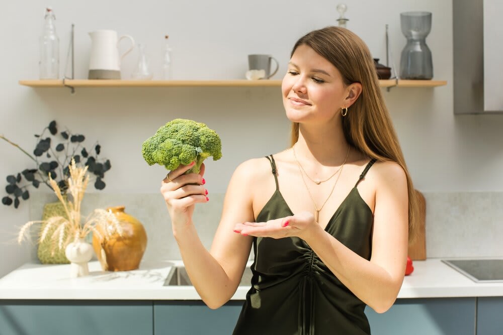 A young woman standing in her kitchen, holding a head of broccoli up in one hand and gesturing towards it with the other.