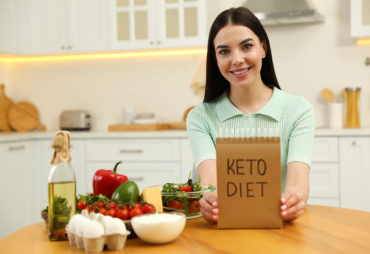 A woman smiling, holding a notebook reading "Keto Diet" with various items on the kitchen counter, such as olive oil, bell peppers, tomatoes, yoghurt, eggs, and avocado.
