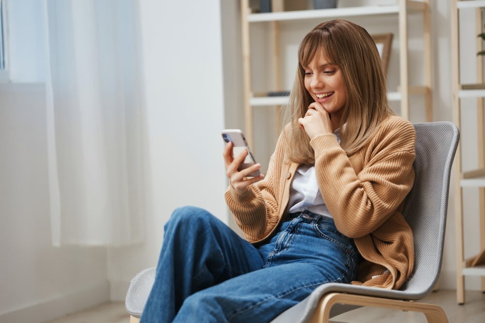 A young woman seated in her living room, smiling at something on her phone screen.