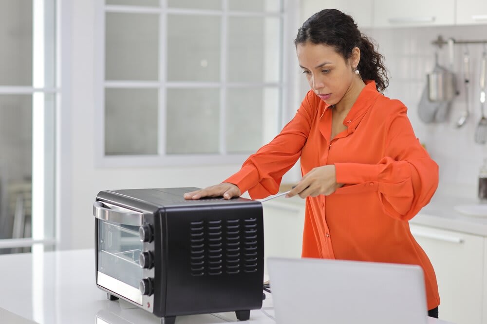 A woman standing in her kitchen, trying to open the back of her microwave with a screwdriver. 
