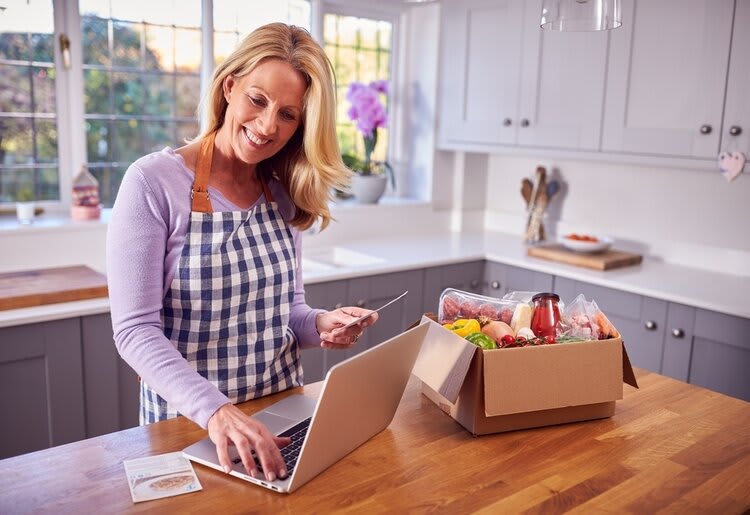 A woman standing in her kitchen with her laptop and a meal kit box on the table.