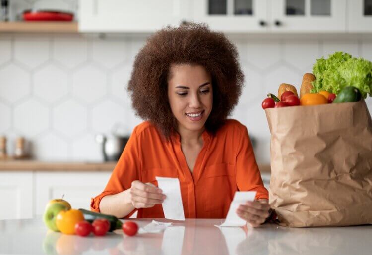 A woman sitting at a table with a bag of groceries comparing orders of delivery services.