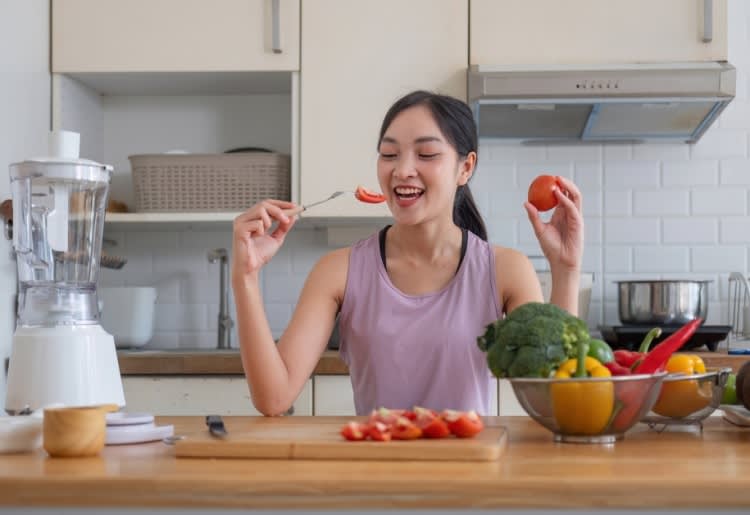 A healthy woman eating a tomato and preparing a low-sugar meal in her kitchen.