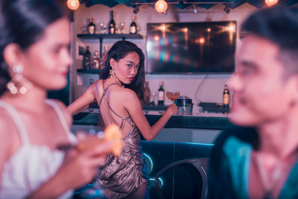 A young woman standing by a bar, staring at a couple in conversation.