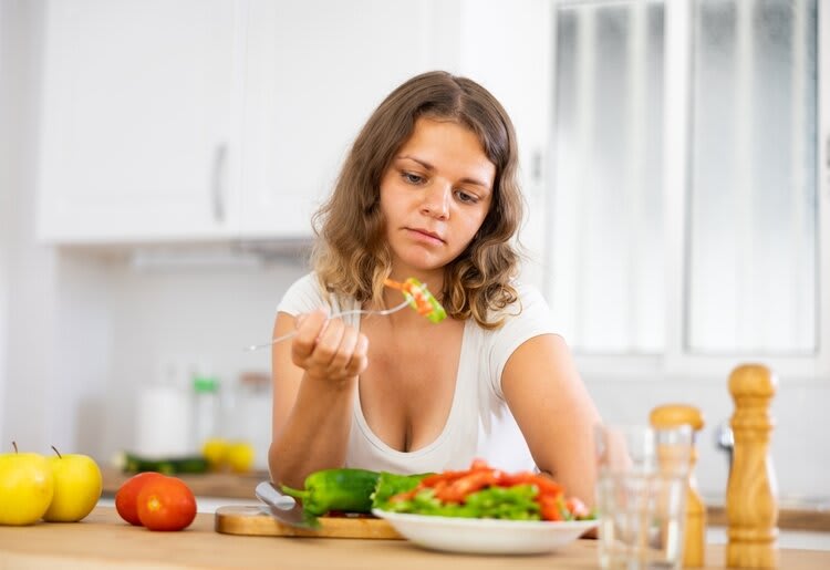 A teenager standing in the kitchen eating a salad, with a discontent expression on her face.