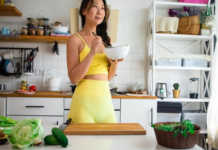 Woman in workout clothes standing in kitchen holding bowl and spoon