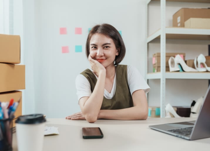 A small business owner with a brick-and-mortar store sitting in front of her laptop and boxes of stock moving her business to an e-commerce store