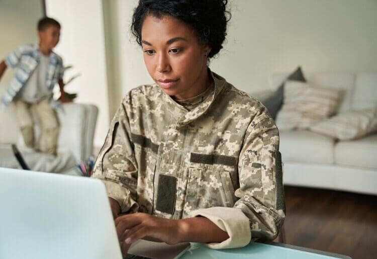 A woman in a military uniform sitting in front of a laptop computer, looking for online trauma therapy services.  