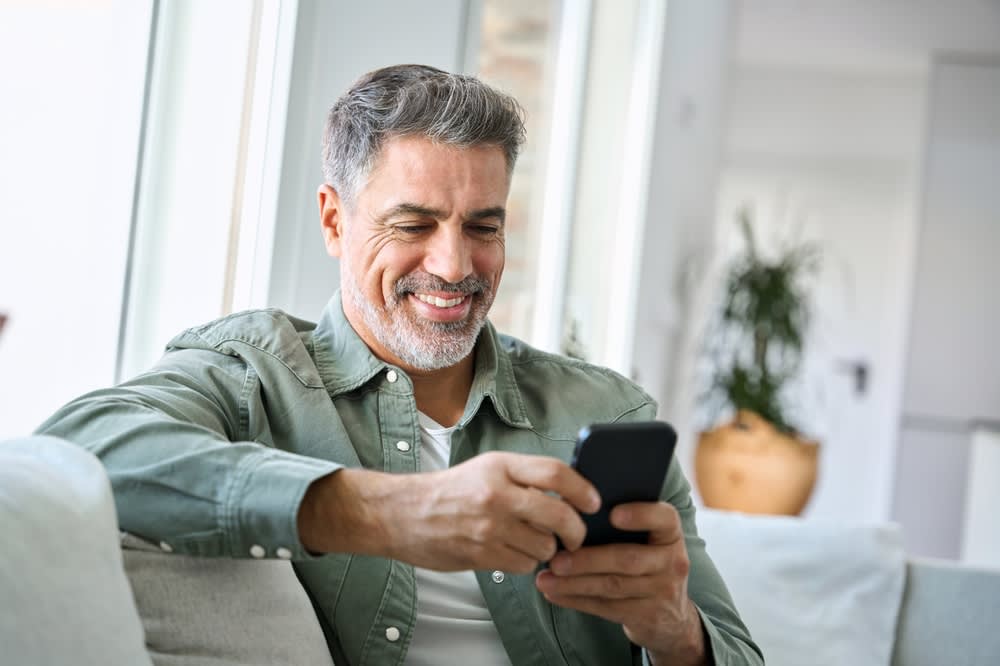 A man over 40 is sitting on a couch looking at his online dating site on his cell phone.