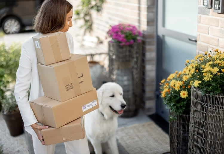 A woman holding a stack of packages and going up her porch.
