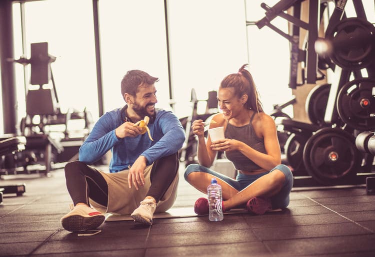 A man and a woman eating post-workout snacks on the floor in a gym.