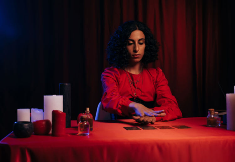A psychic dressed in red, seated at a table featuring various candles, hovering her hand over a row of oracle cards.