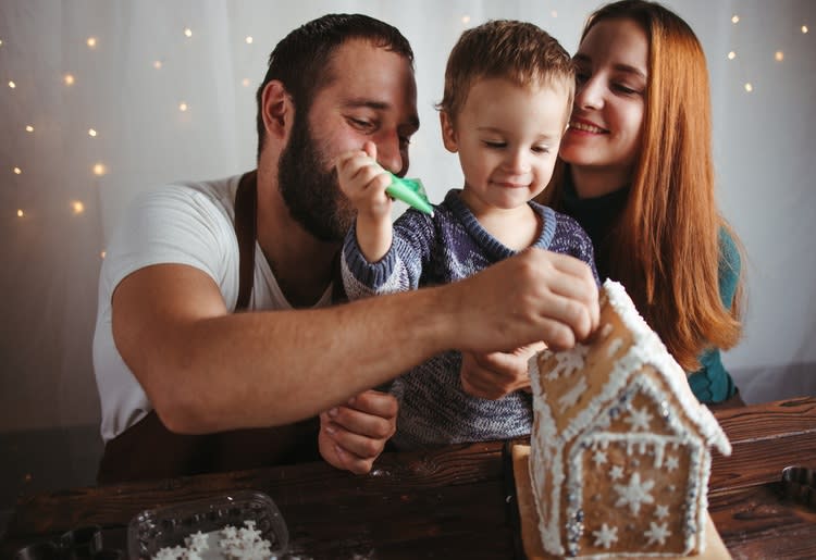 Family of three decorating a gingerbread house together.