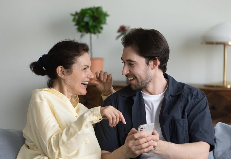 A couple of differing ages (an older woman with dark hair, wearing a light yellow long sleeve shirt and a younger man with dark hair wearing a white t-shirt and dark short-sleeve button-up) laughing together as he shows her something on her phone. They are seated on a couch and a cabinet with two plants, as well as a lamp are visible in the background.