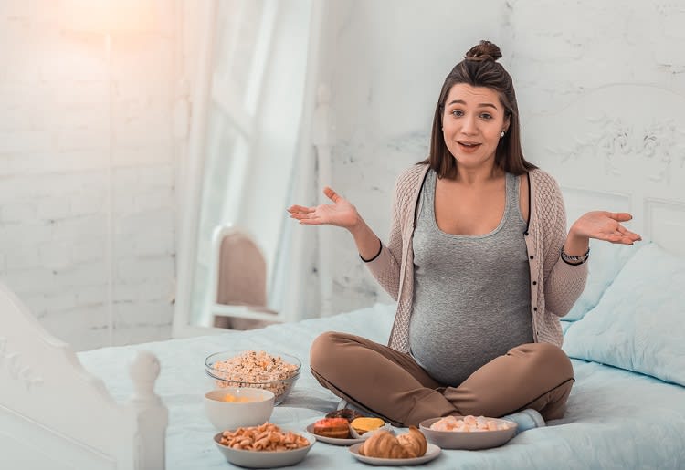 Pregnant woman sitting cross-legged on bed surrounded by bowls of food