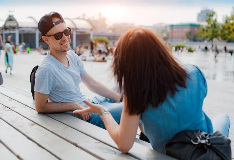 A young man and woman sitting on a bench talking to one another happily