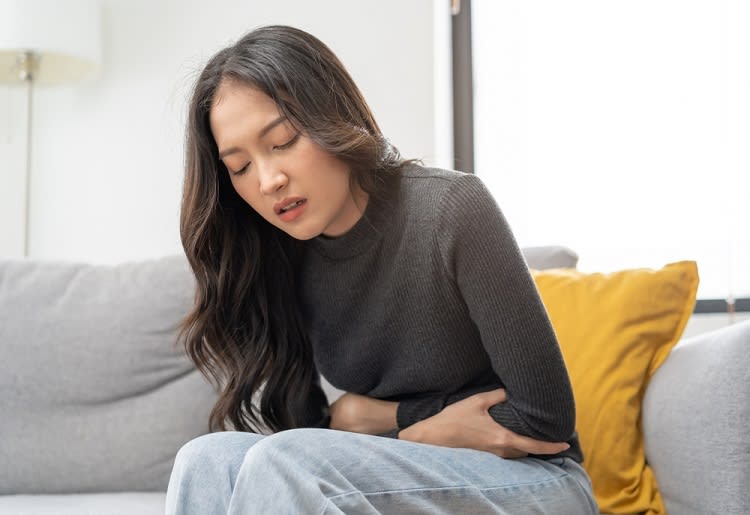 A young woman sitting on her couch while holding her stomach  in discomfort
