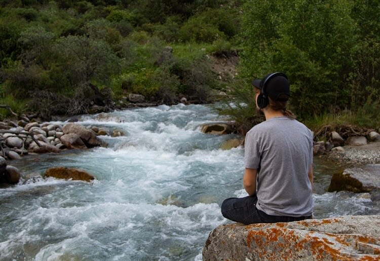 A young man with headphones on sitting on a rock with a flowing river below