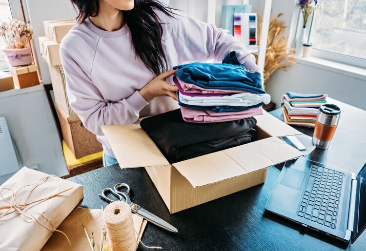A woman wearing a lilac jumper is packing a pile of colorful, folded clothing items into a box, with tied packages, twine, scissors, a thermos mug, and a laptop on the table, with more boxes and dried flowers in the background of a light-filled room