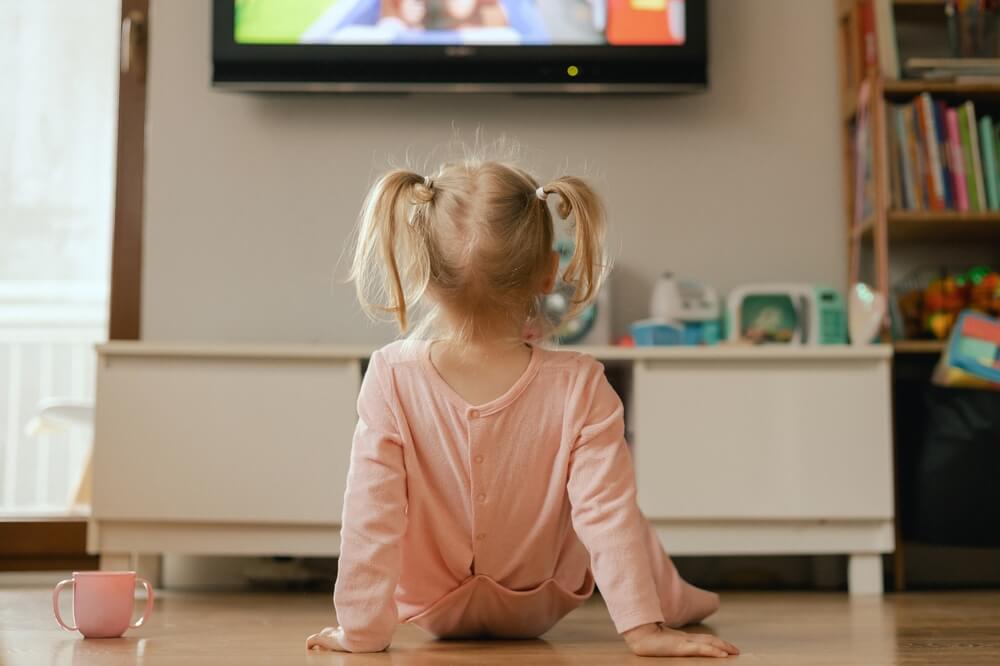 Portrait of happy baby playing with toys and watches cartoons on TV in playroom.