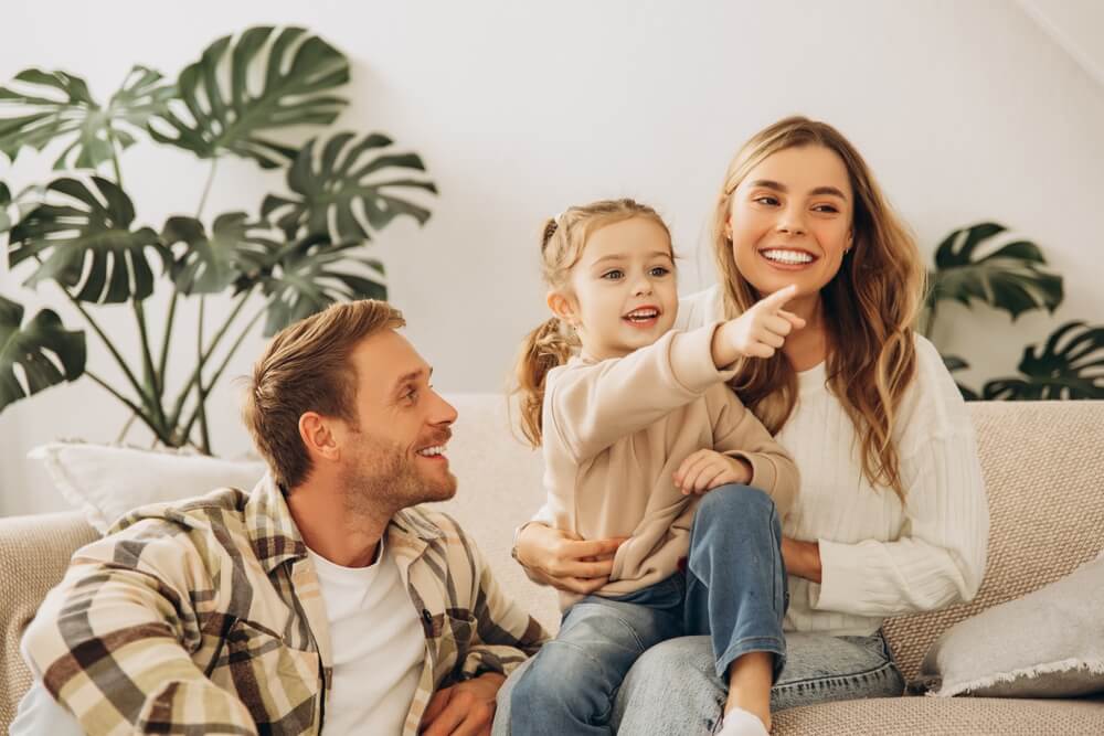 Portrait of happy family, smiling mother and father embracing together, little girl pointing finger, showing, sitting on comfortable sofa in living room at home. 